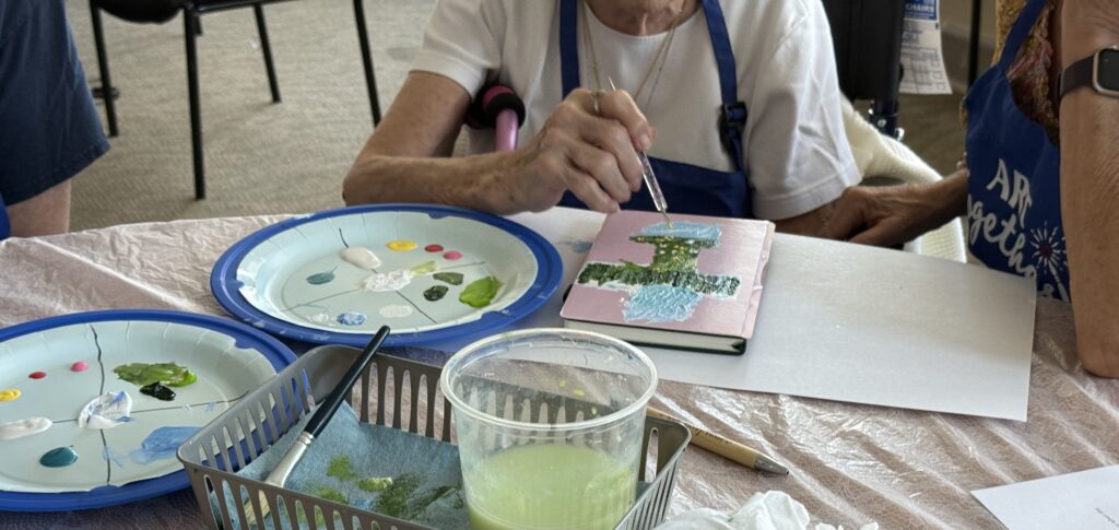 Older adult painting journal cover with a landscape assisted by a volunteer